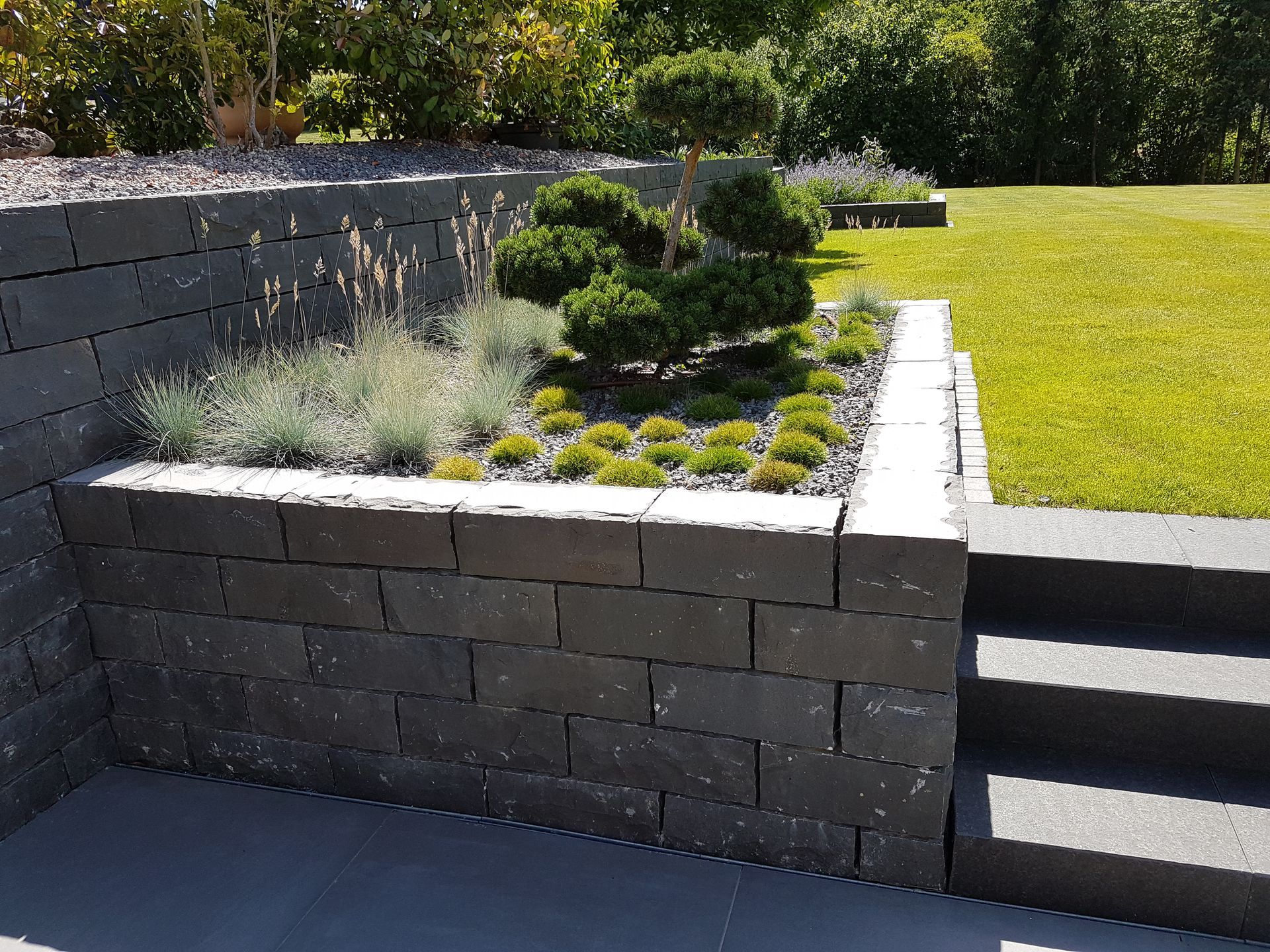 Ein Hochbeet mit dunklen Ziegelmauern, in dem Pflanzen und Sträucher wachsen, grenzt an eine Rasenfläche.