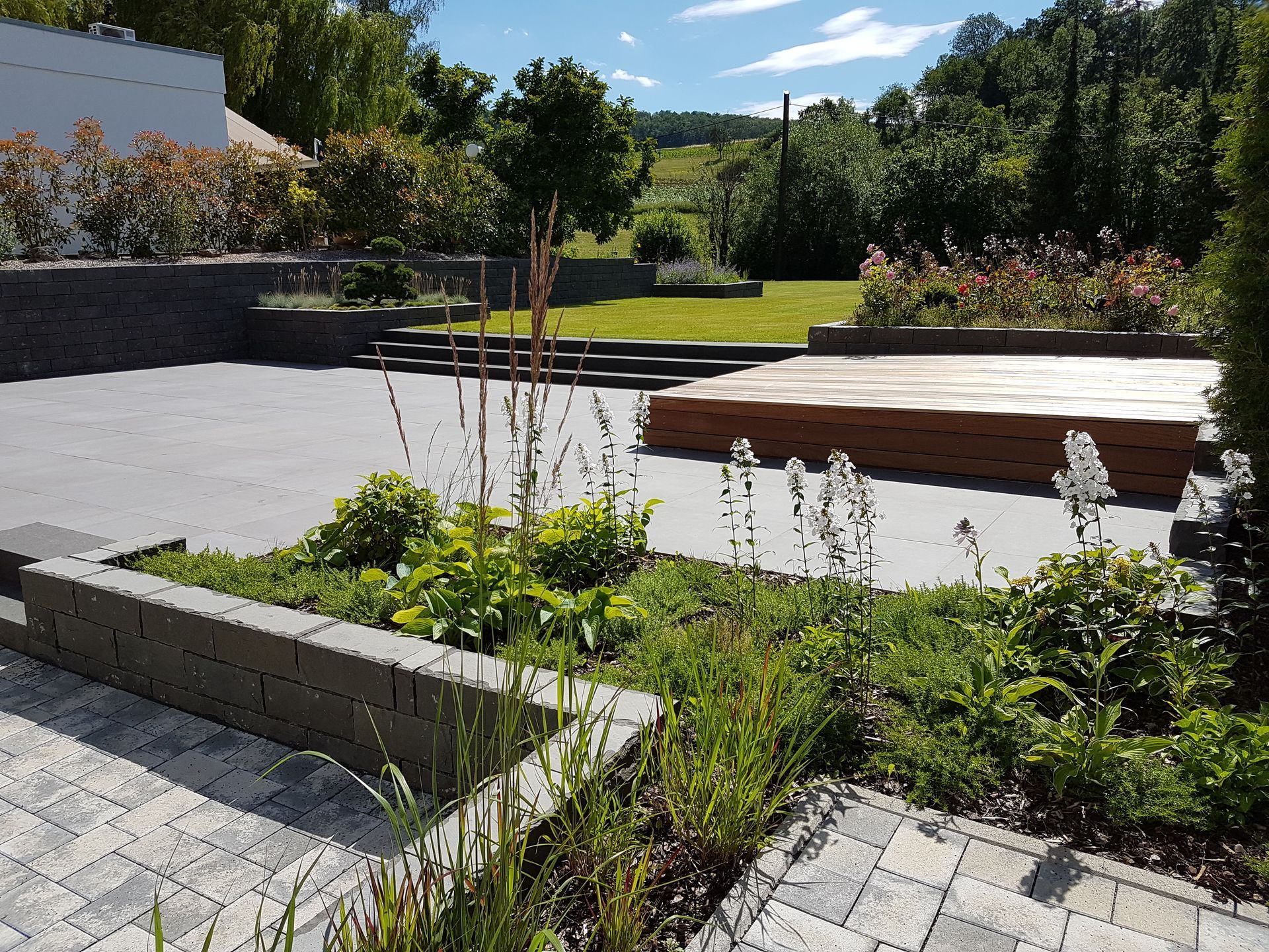 Ein landschaftlich gestalteter Hinterhof mit Terrasse, Holzdeck und üppigen grünen Pflanzen unter einem sonnigen Himmel.