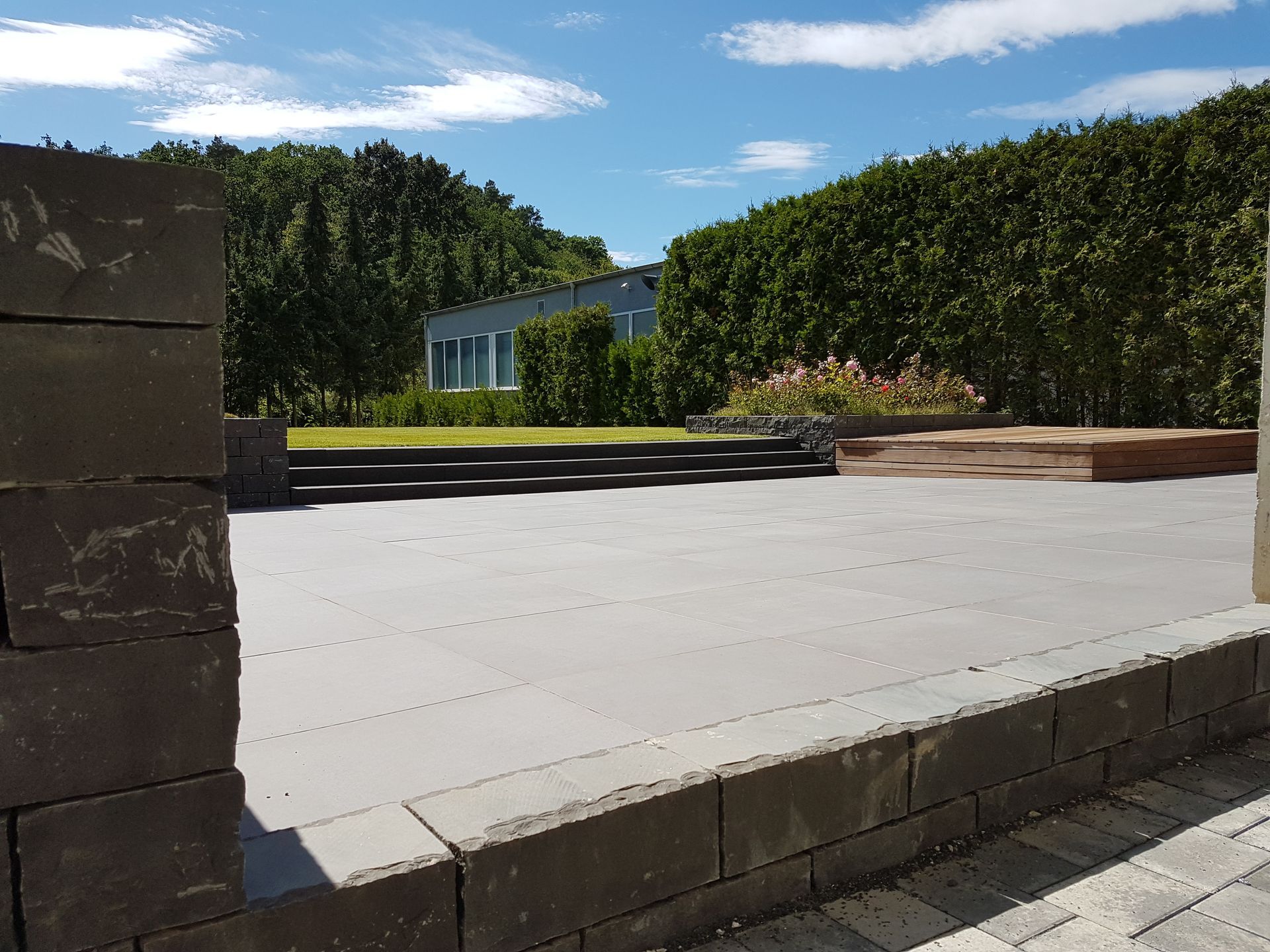 Steinterrasse mit dunklen Backsteinmauern, Rasen und einem kleinen Gebäude im Hintergrund. Blauer Himmel.
