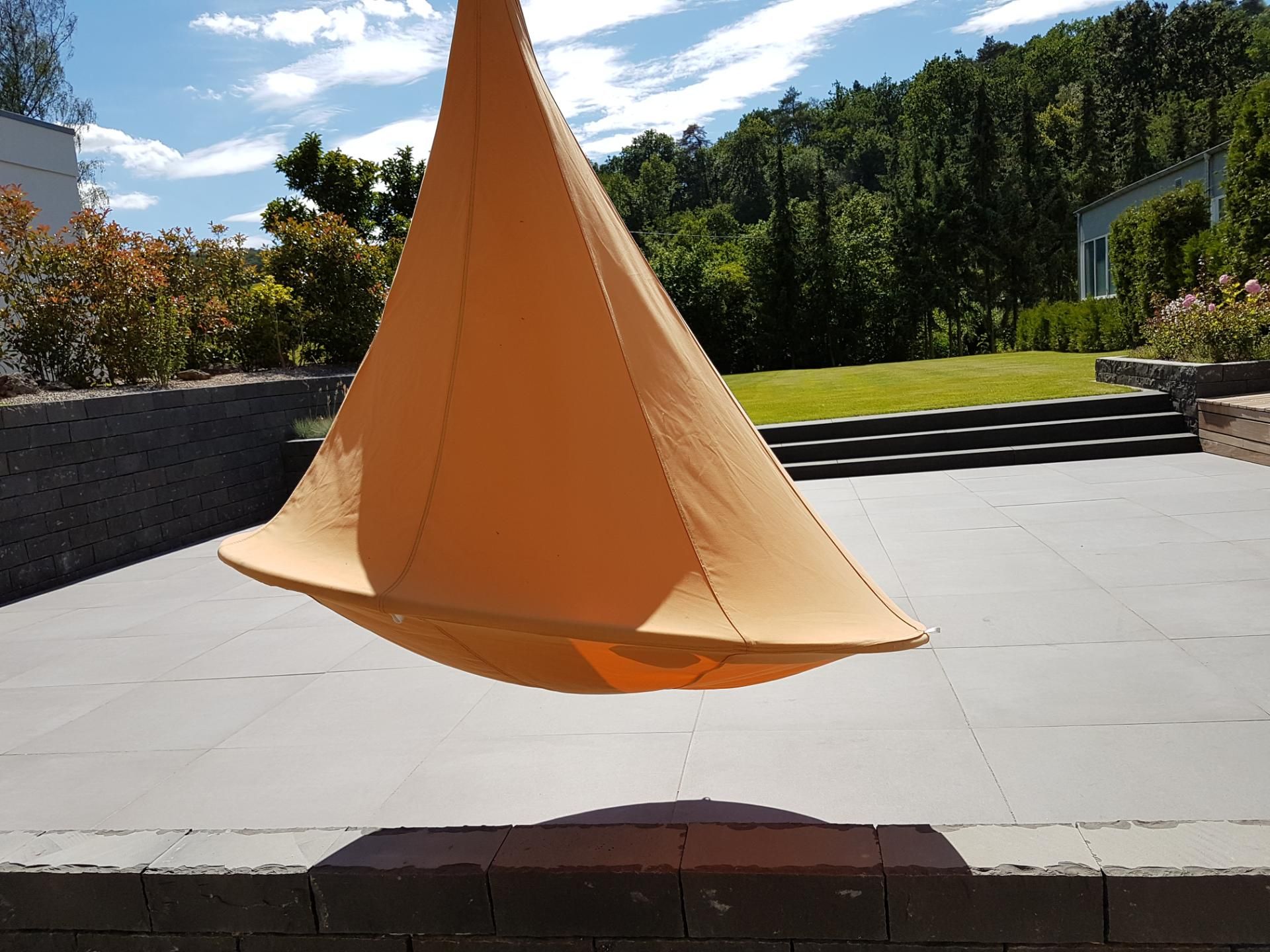Orangefarbener Hängesessel über einer grauen Terrasse mit grünem Rasen und Bäumen im Hintergrund.