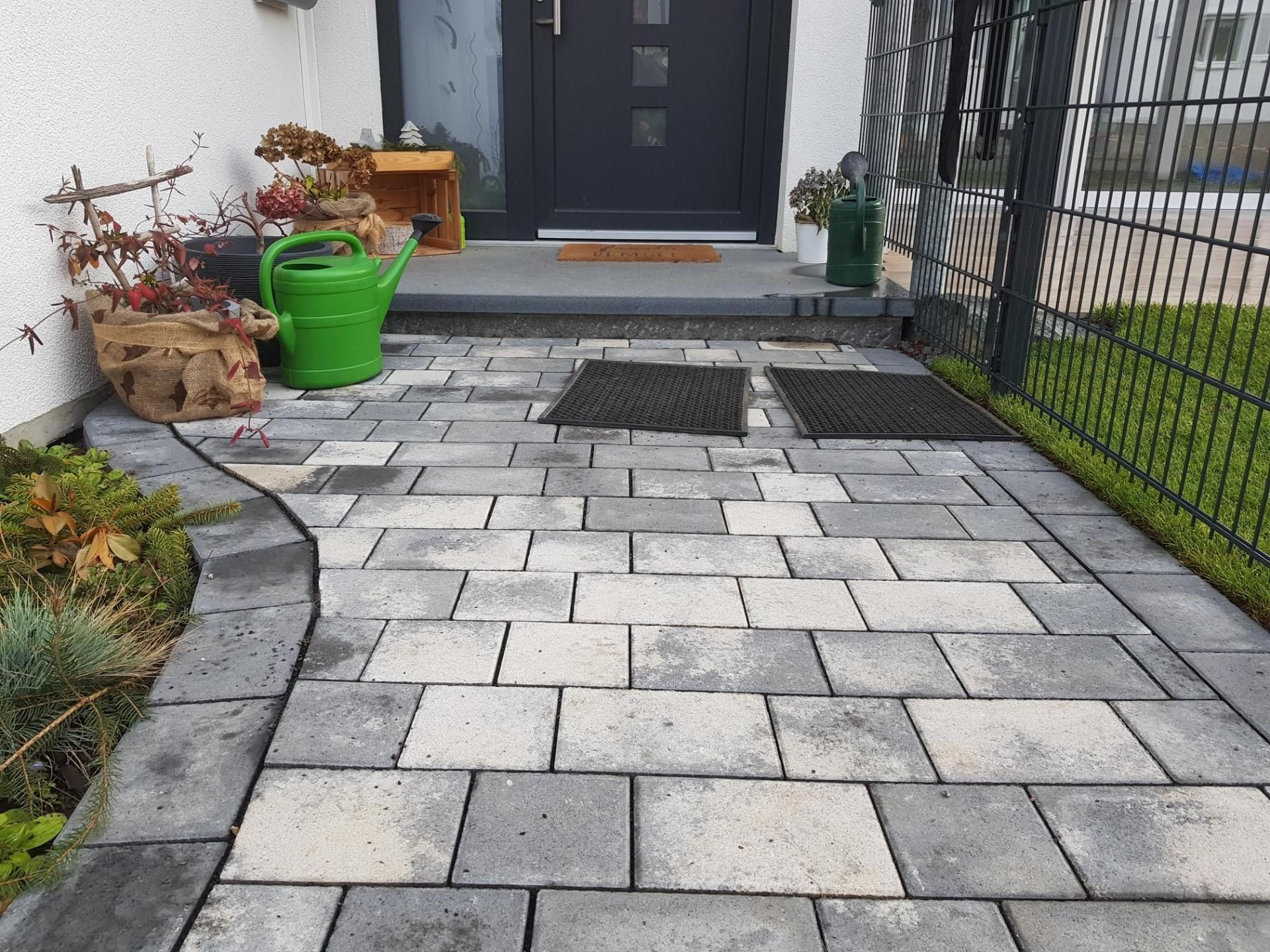Ziegelweg, der zu einer grauen Haustür mit einer grünen Gießkanne und Pflanzen führt.