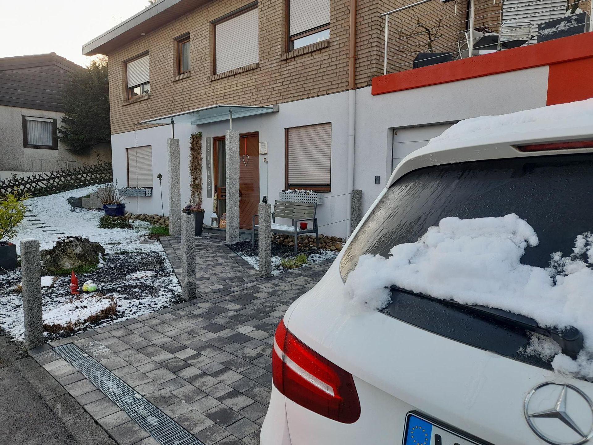 Schneebedecktes Haus mit einem davor geparkten weißen Auto; grauer Gehweg und Vorgarten.