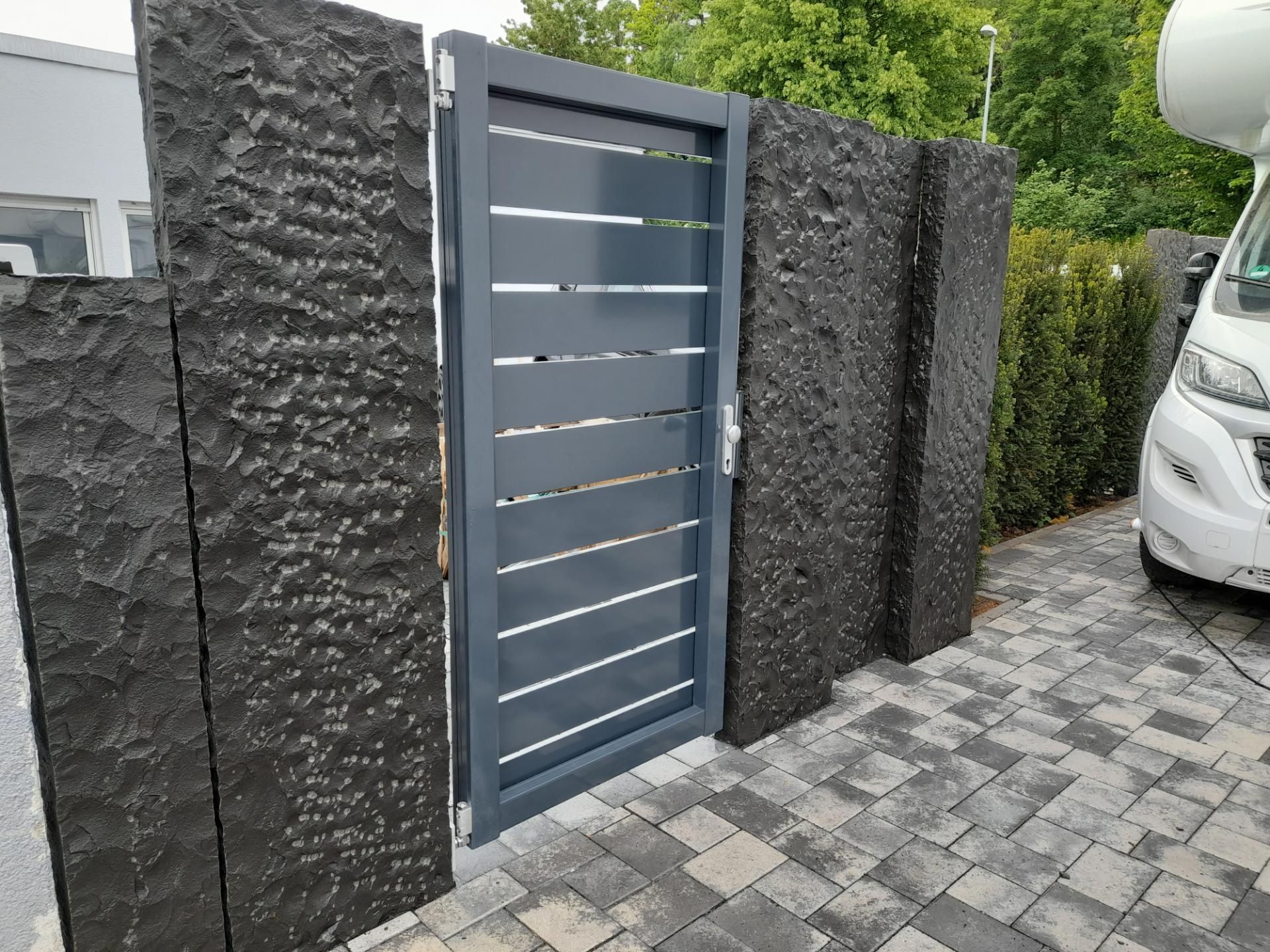 Graues Tor mit horizontalen Lamellen zwischen schwarzen strukturierten Steinsäulen auf einer gepflasterten Auffahrt.
