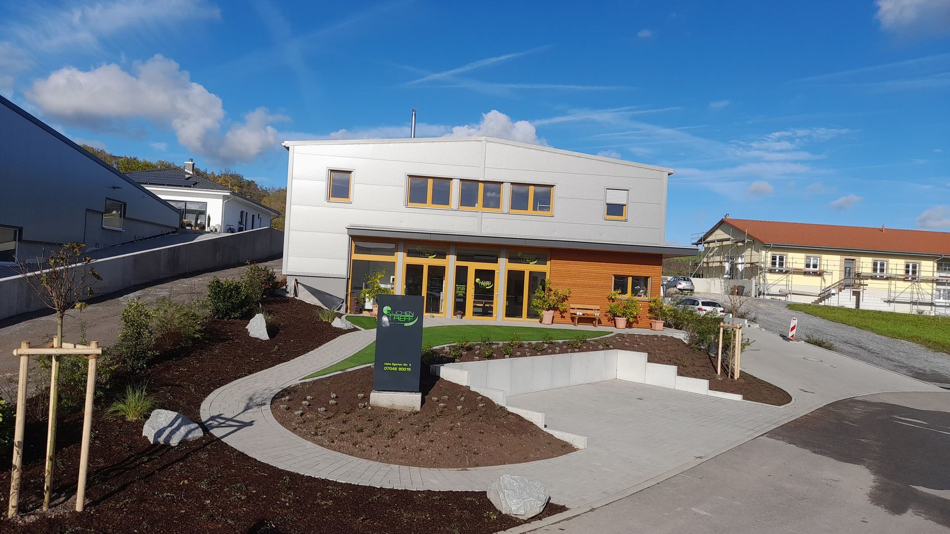 Modernes Gebäude mit silberner Fassade und hölzernem Eingangsbereich, umgeben von einer gepflegten Landschaft und strahlend blauem Himmel.