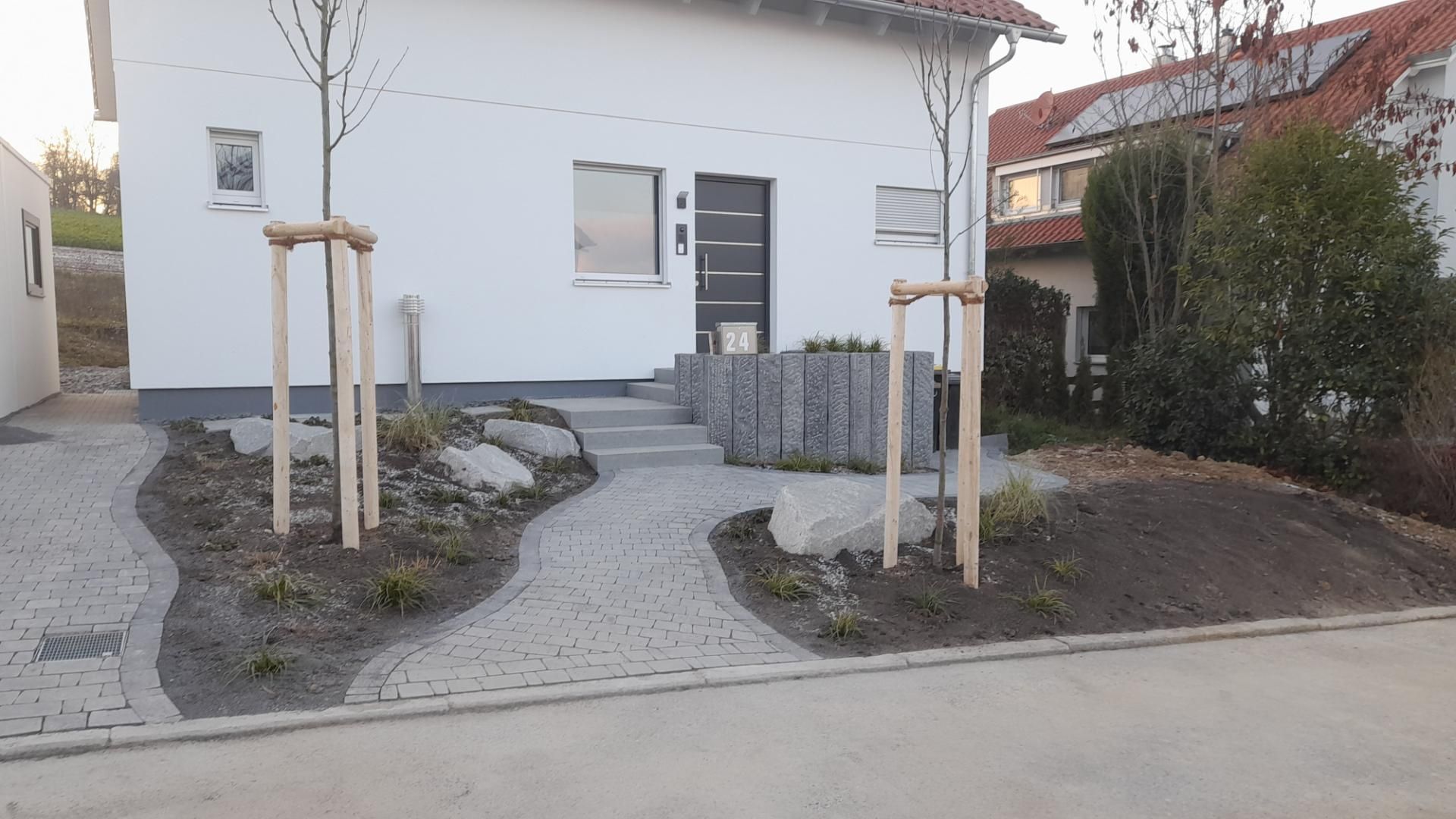Weißes Haus mit Steinweg und Landschaftsgestaltung; davor stehen zwei junge Bäume mit Stützen.