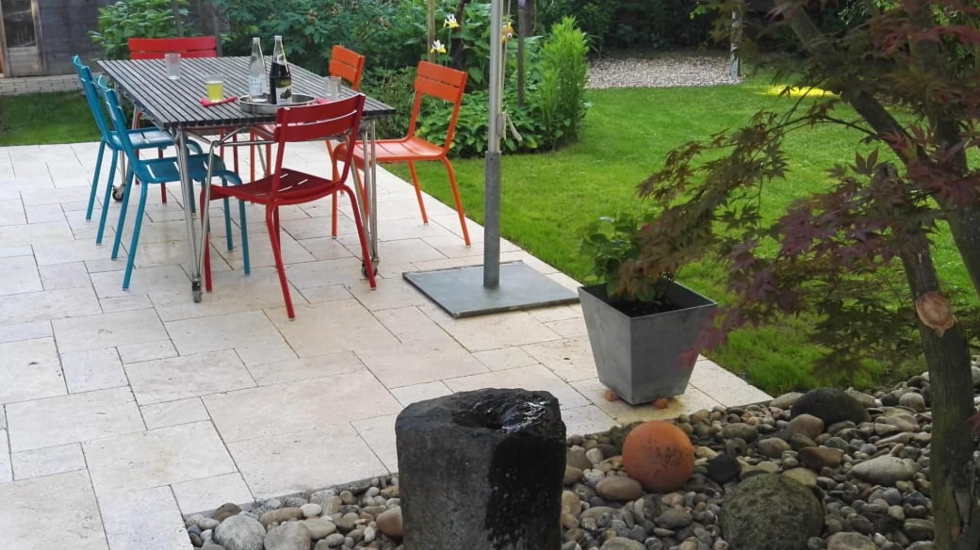 Essbereich im Freien mit einem Tisch, bunten Stühlen und einem Steinbrunnen.