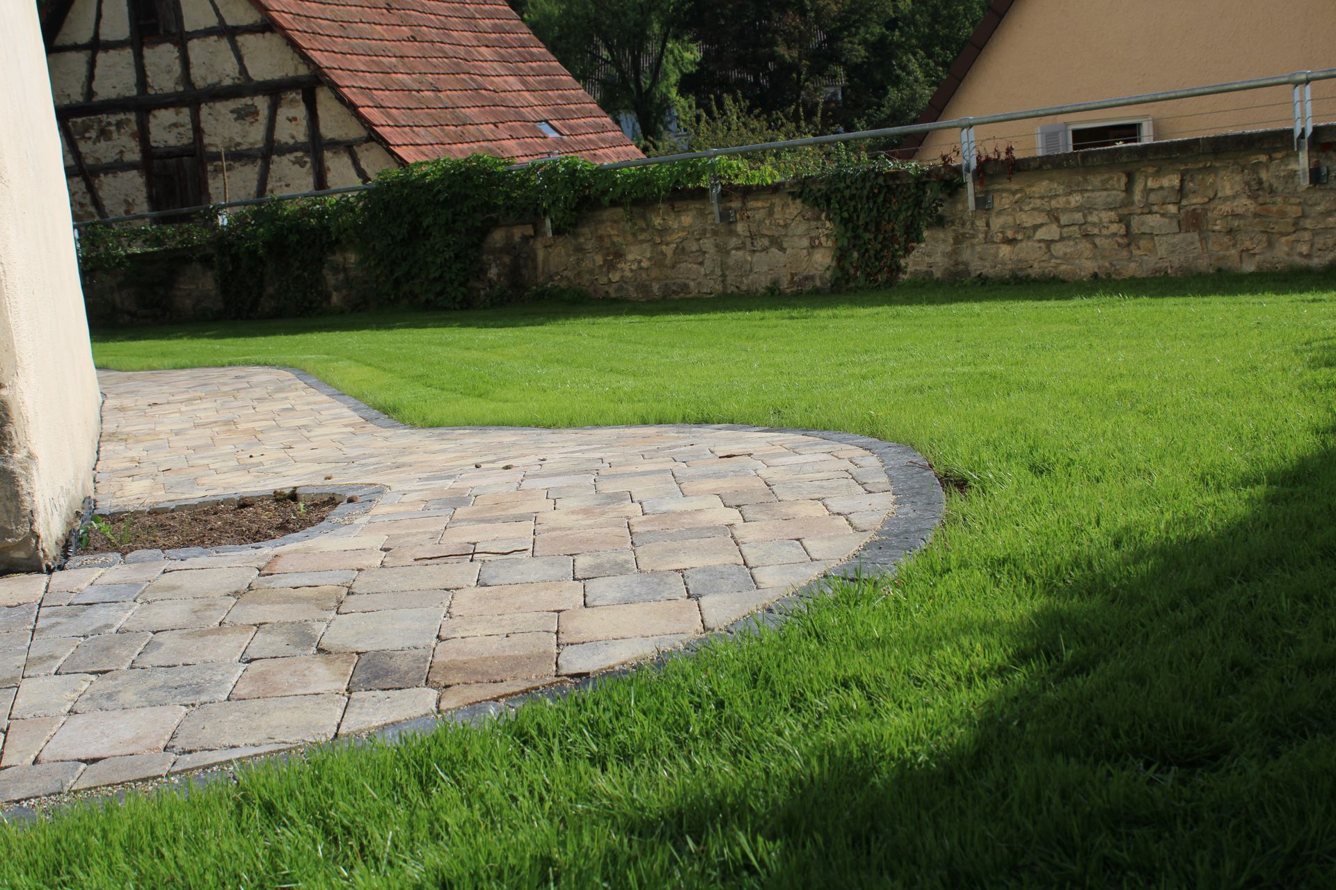 Eine gepflasterte Terrasse geht in eine Rasenfläche über, im Hintergrund sind eine Steinmauer und ein Fachwerkhaus zu sehen.