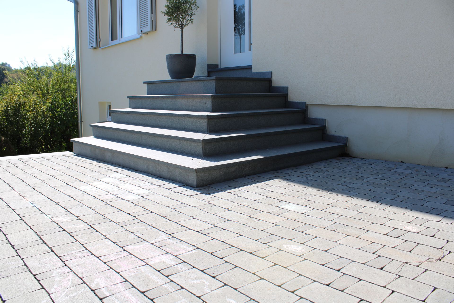Steintreppe führt zu einem beigefarbenen Hauseingang. Gepflasterte Terrasse im Vordergrund. Olivenbaum im Topf.