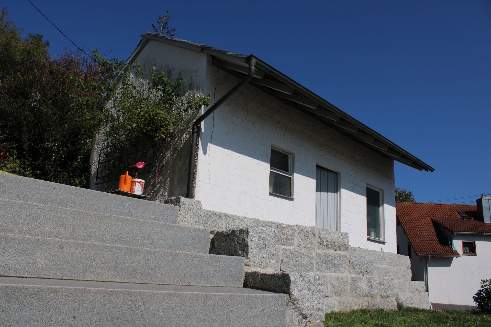 Weißes Gebäude mit Steintreppe, in der Ferne ein Haus mit rotem Dach, blauer Himmel.