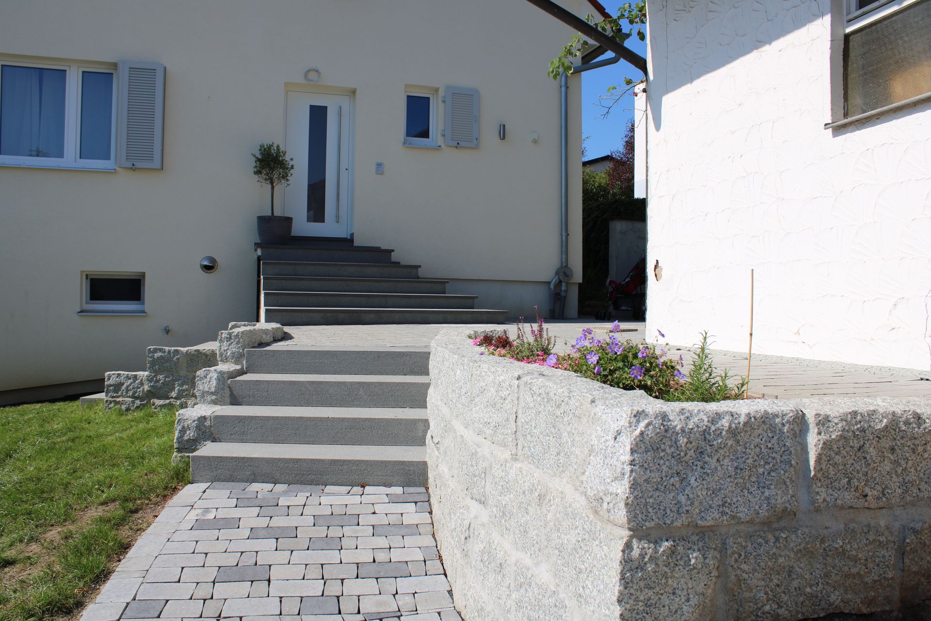 Beiges Haus mit grauen Steinstufen und Stützmauer; davor Blumen und ein Gehweg.