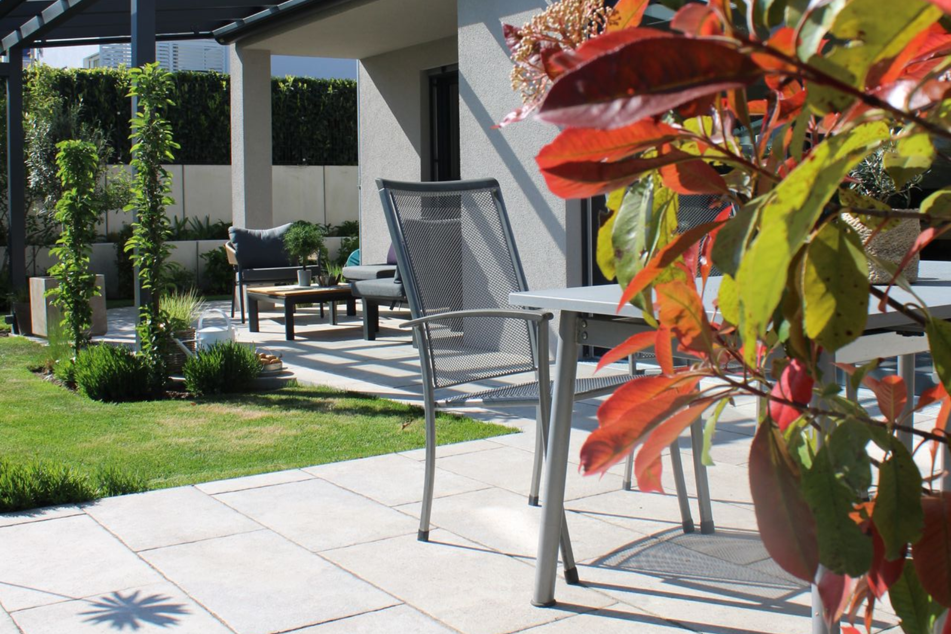 Terrasse mit Metalltisch und -stühlen, üppiges Grün, Hausfassade, sonniger Tag.