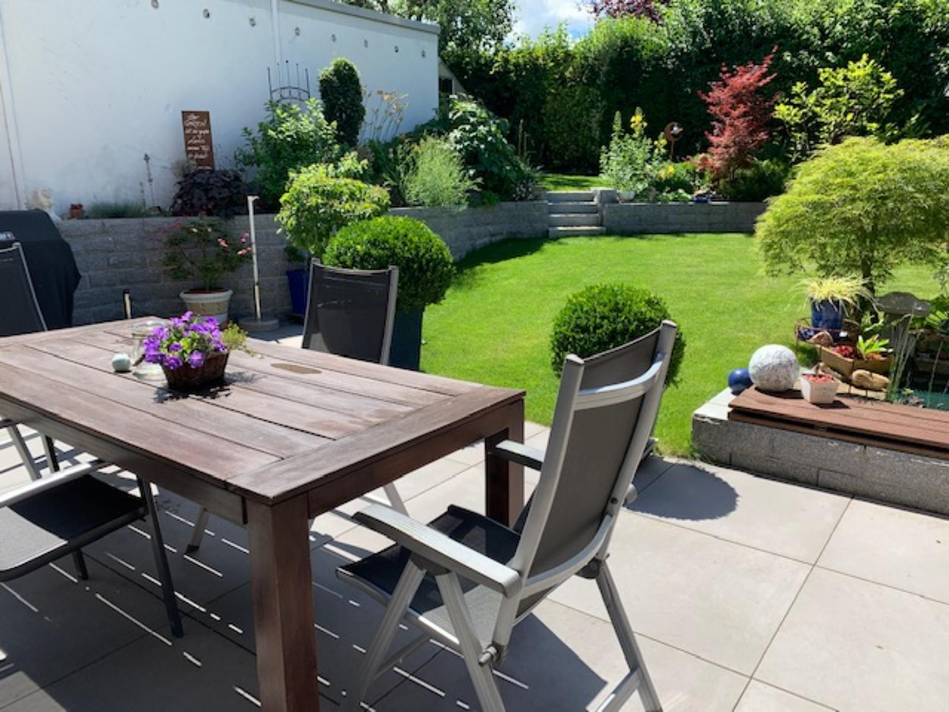 Terrasse mit Holztisch und Stühlen mit Blick auf einen grünen Rasen und Garten mit leuchtenden Pflanzen.