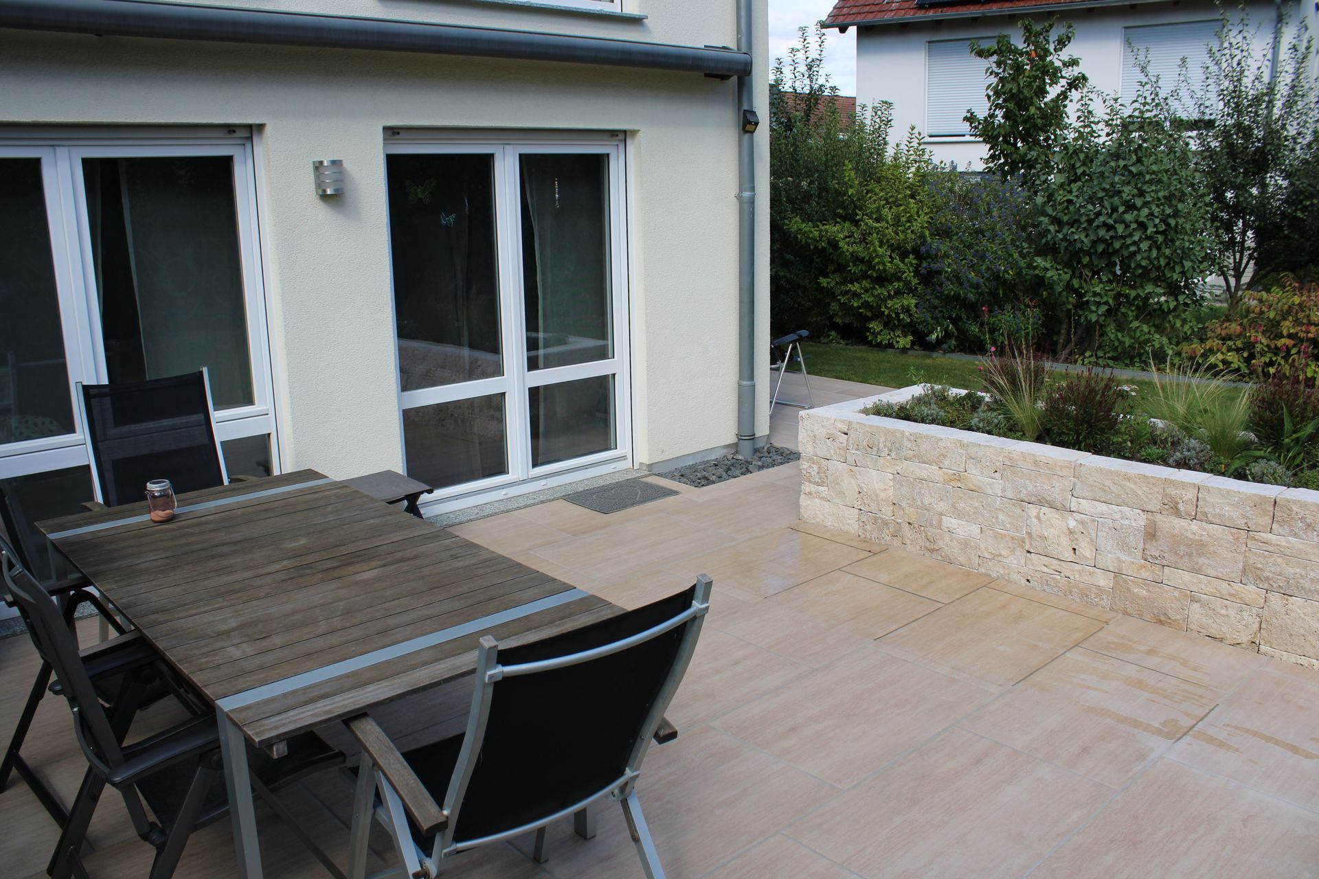 Eine Stützmauer aus Stein grenzt an eine gepflasterte Terrasse neben einem weißen Haus mit Weinreben und Grünpflanzen.