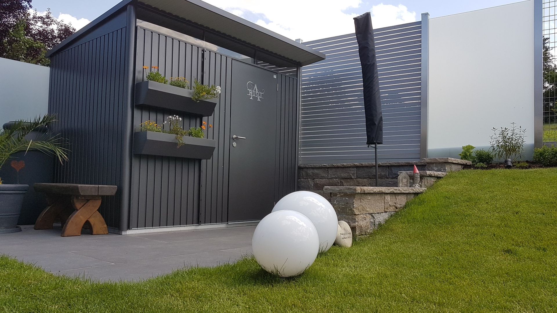 Moderner Schuppen mit dunkelgrauer Verkleidung, Pflanzkästen und dekorativen weißen Kugelleuchten auf dem Rasen.