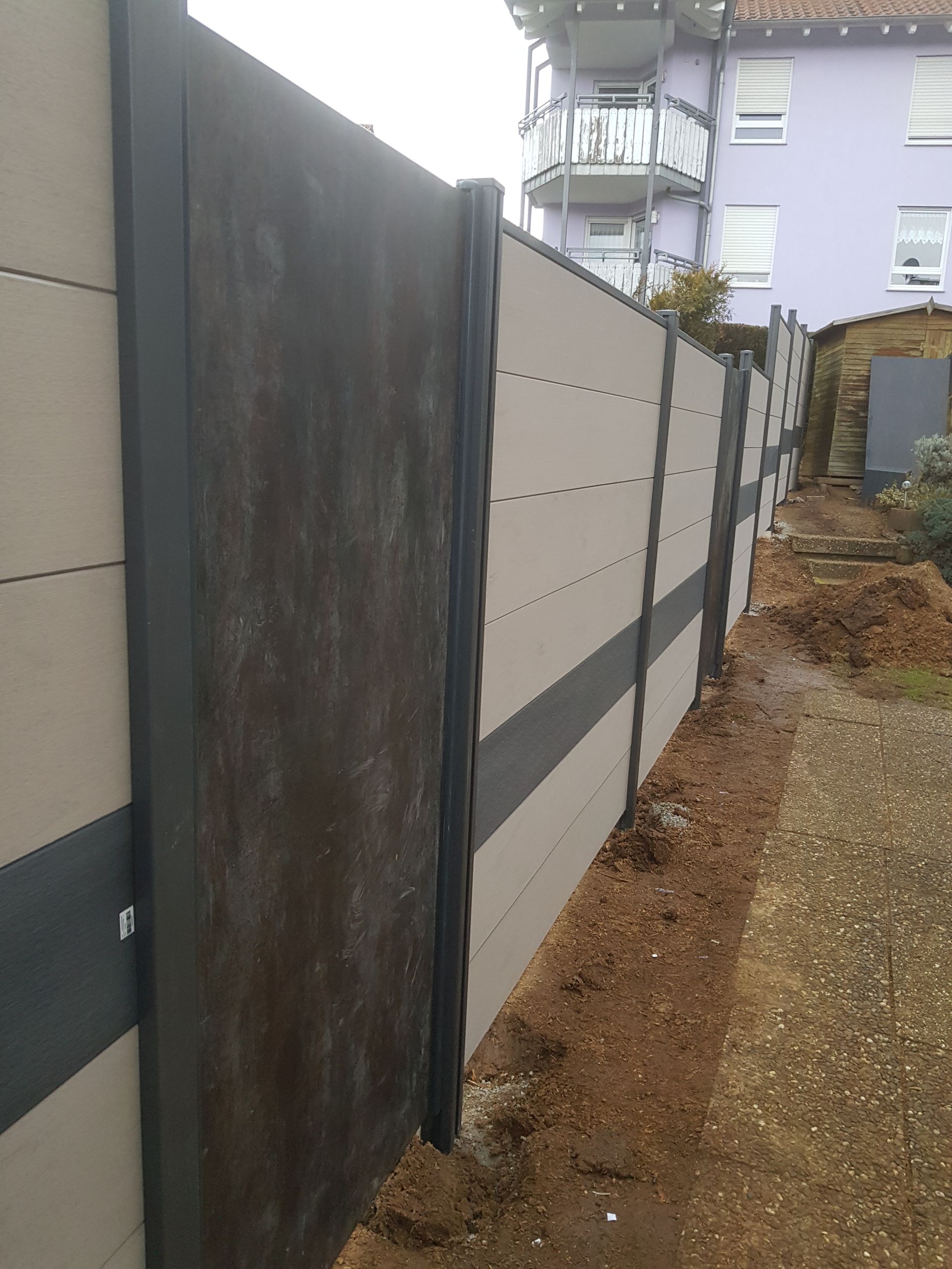 Beige-schwarzer Betonzaun entlang eines Feldwegs, im Hintergrund ein violettes Gebäude.