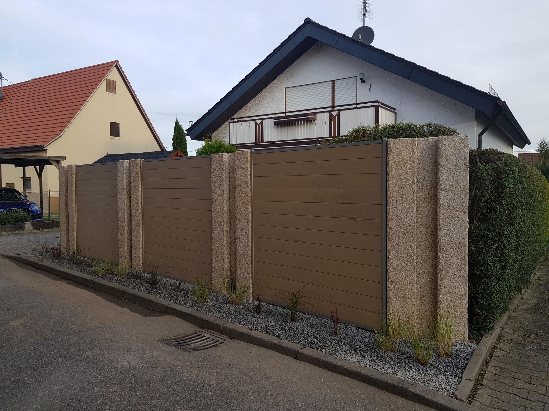 Vor einem Haus befindet sich ein brauner Steinzaun. Im Hintergrund ist ein Kiesweg mit einem hellbraunen Gebäude und rotem Dach zu sehen.