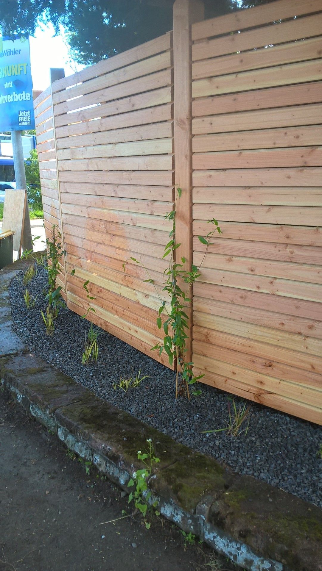 Ein Holzzaun mit horizontalen Brettern neben einem dunklen Felsbett und einer verwitterten Betonbordsteinkante, auf der einige Pflanzen wachsen.