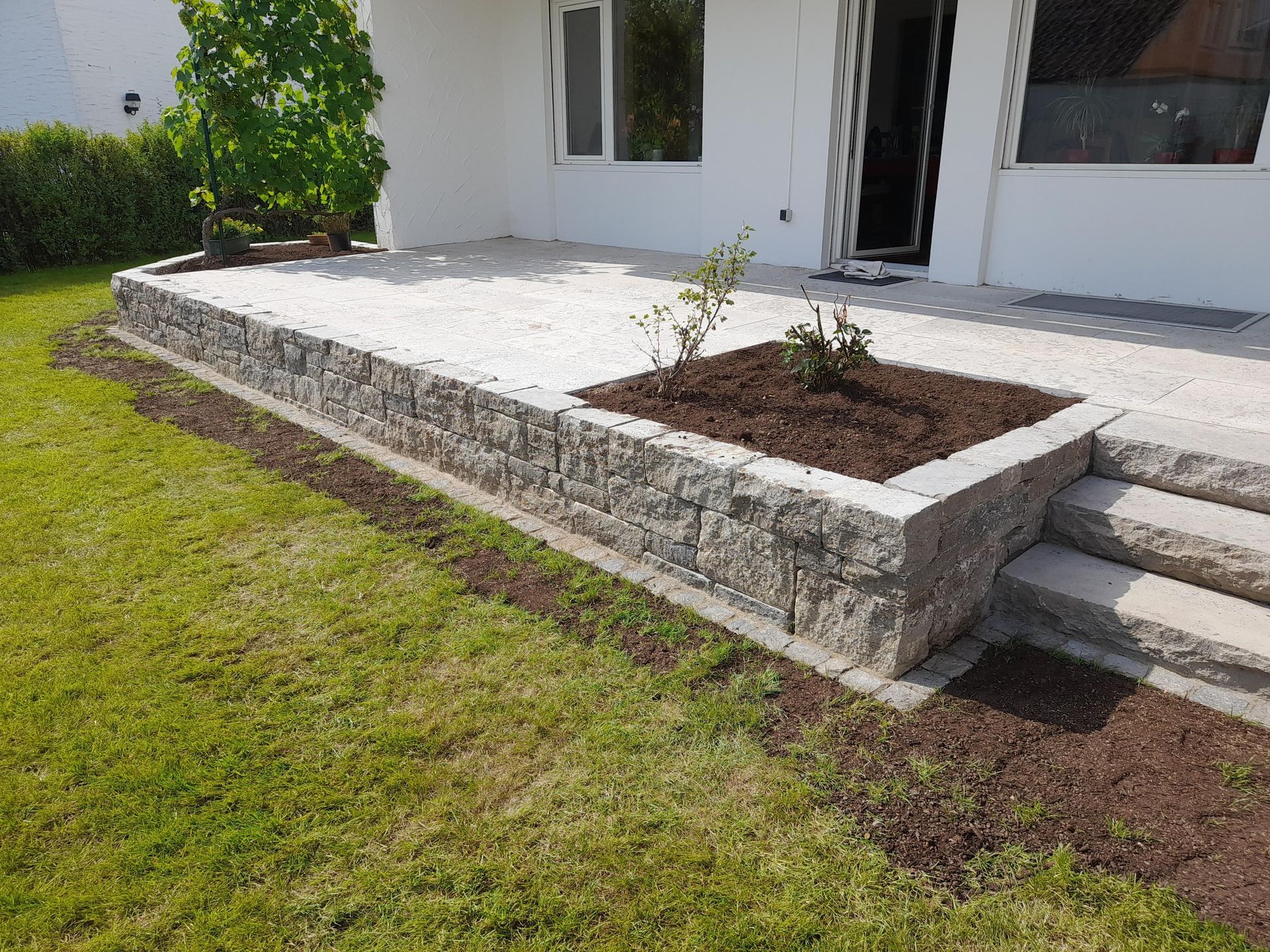Steinterrasse mit Pflanzkasten und Stufen, angrenzend an einen Rasengarten.