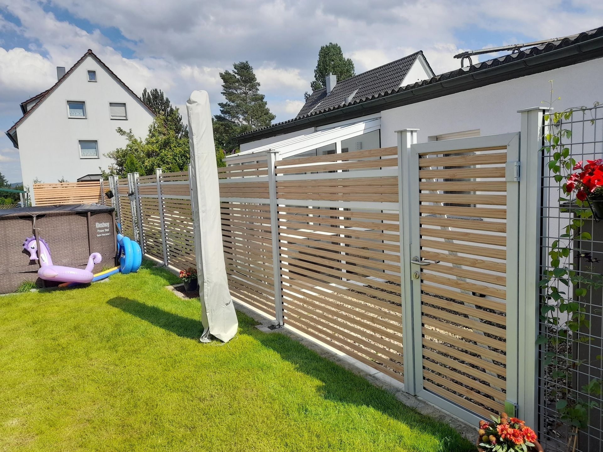 Ein Zaun aus Holzlatten und Metall umgibt eine grüne Rasenfläche, im Hintergrund ist ein Haus zu sehen.
