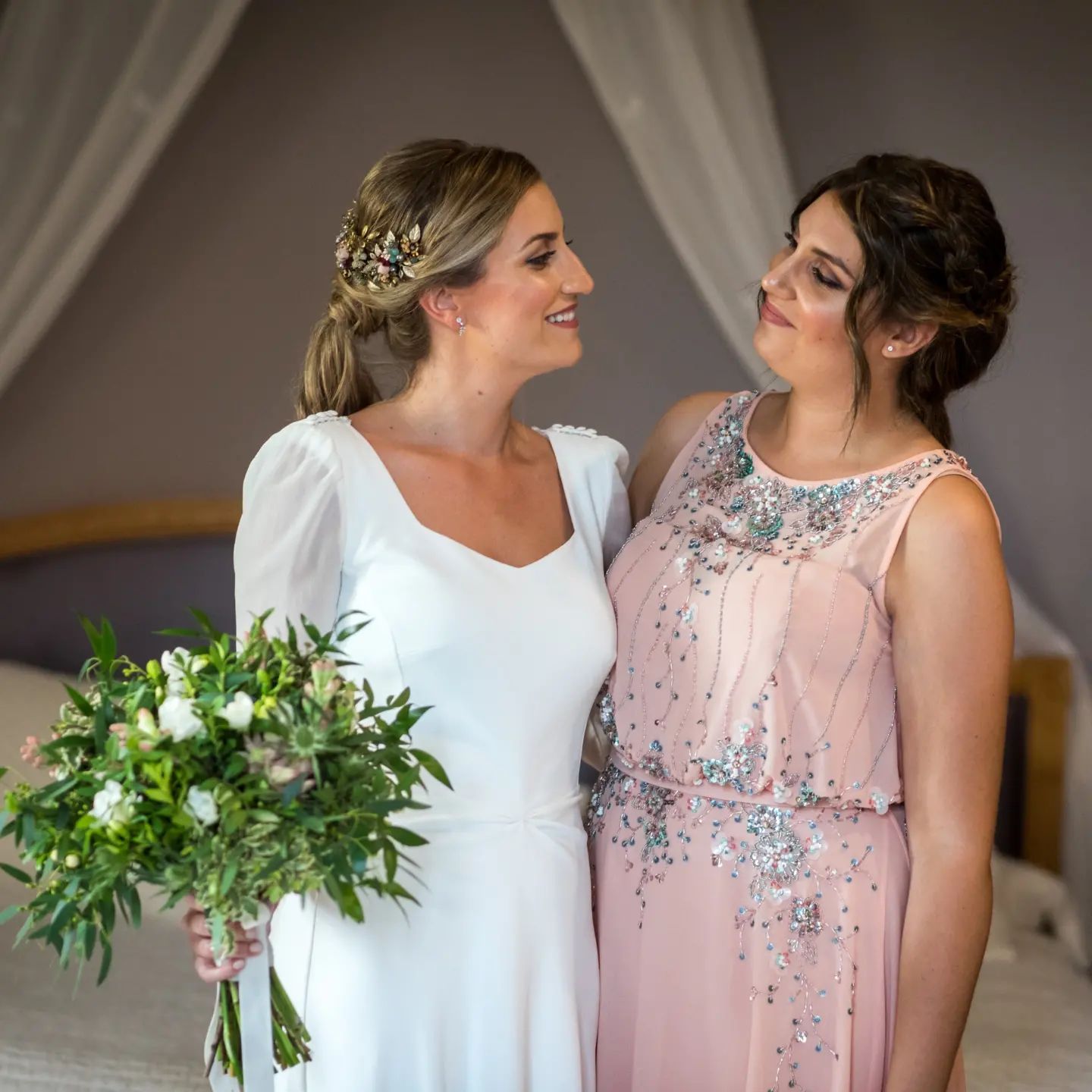 Una novia y su dama de honor posan para una fotografía en un dormitorio.