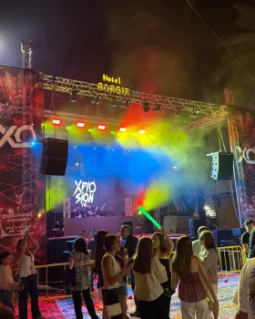 Escenario de conciertos con luces de colores, artistas y público disfrutando de la música. 