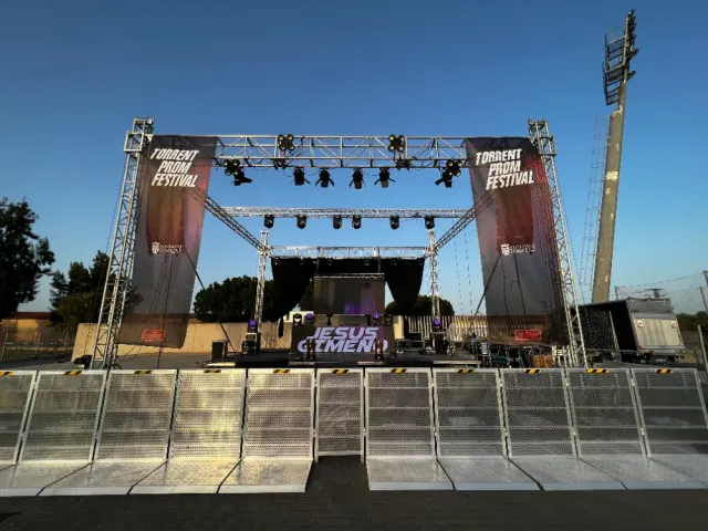 Escenario preparado para un concierto. Incluye iluminación, pancartas y barreras. Cielo azul.