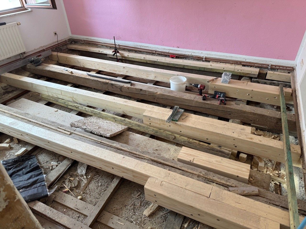 Holzbalken auf einer offenen Bodenbaustelle in einem Raum mit rosa Wand. Der Boden wurde entfernt und wird neu aufgebaut.