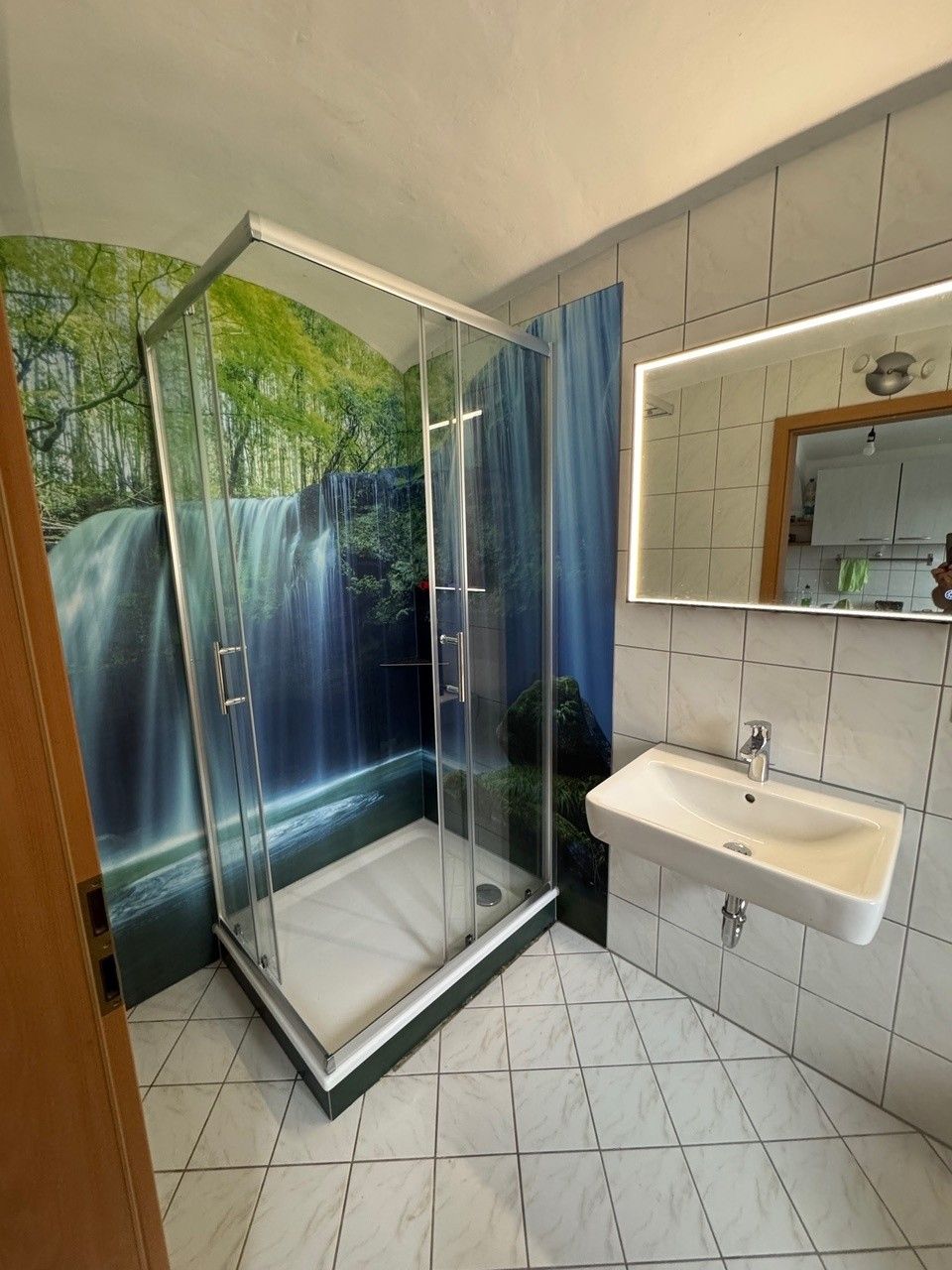 Badezimmer mit gefliestem Boden, Eckdusche mit Naturmotiv an den Wänden und Waschbecken.