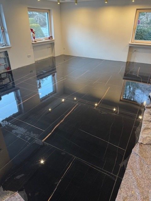 Ein Fliesenleger verlegt eine Wandfliese. Er drückt sie mit einer Hand an, während er auf dem Boden kniet, umgeben von neuen Fliesen und Kleber.