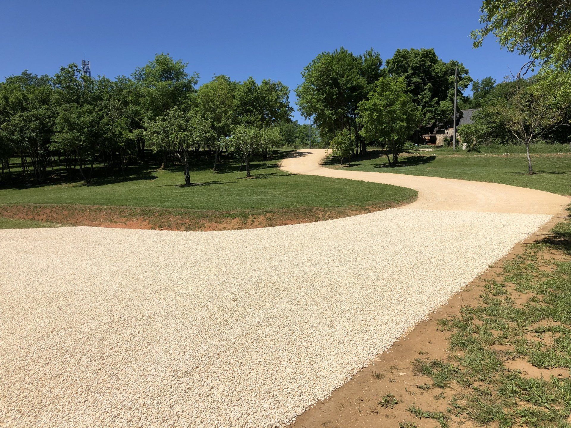 Chemin de gravier sinueux traversant un parc verdoyant et ensoleillé, avec des arbres et un ciel bleu.