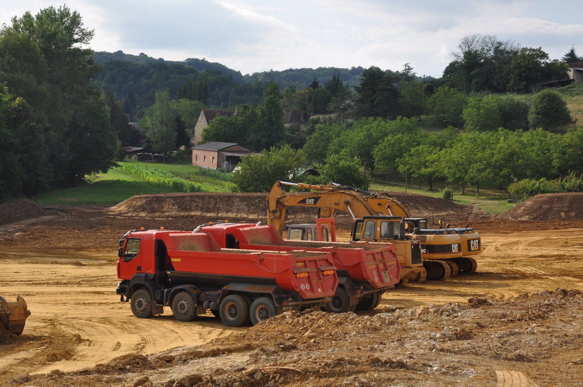 Un camion-benne rouge et une pelleteuse jaune sur un chantier boueux, avec des arbres et des collines en arrière-plan.
