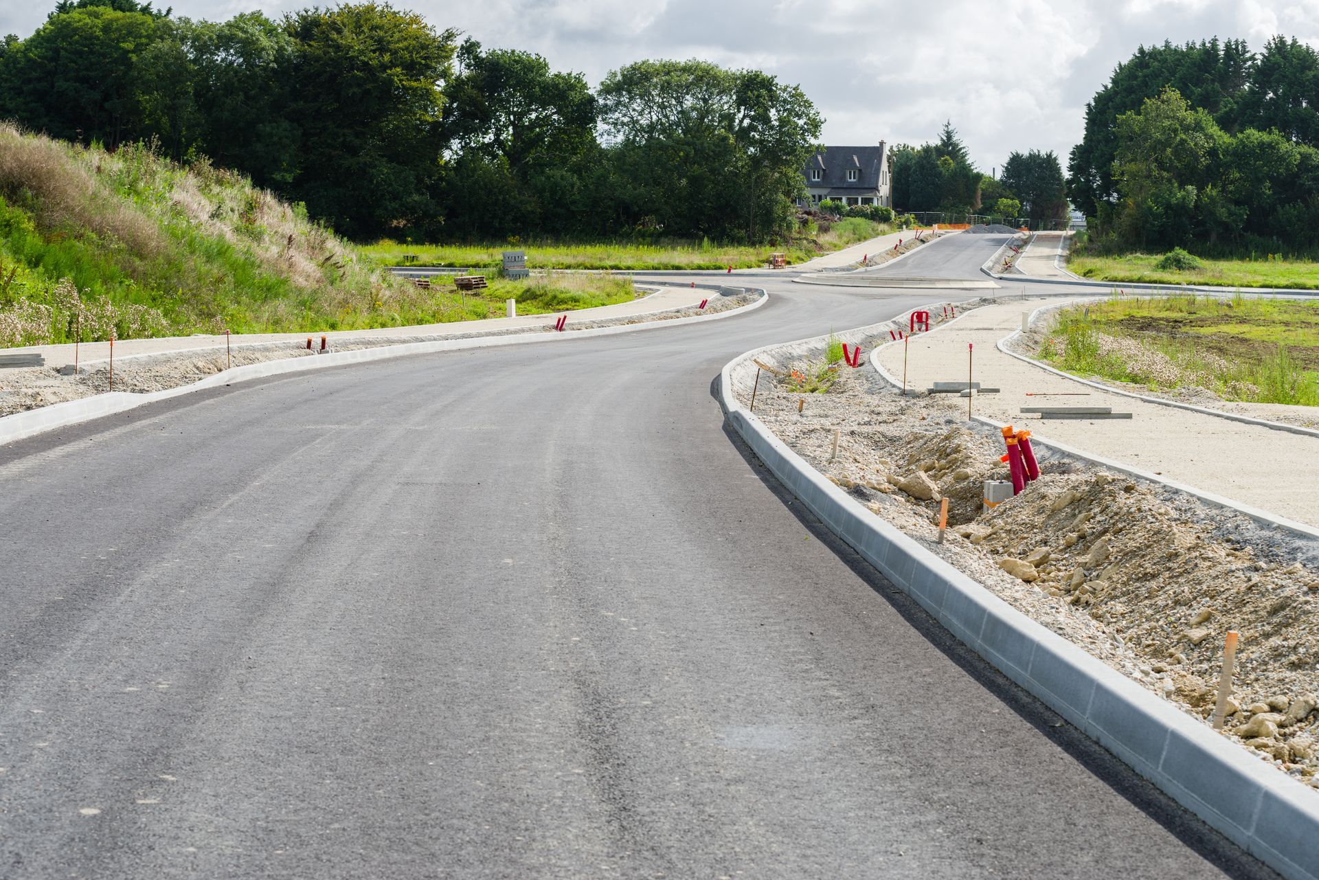 Route sinueuse nouvellement asphaltée en construction, balisée par des cônes et bordée d'arbres, avec accotements en gravier et jalonnés de pierres.