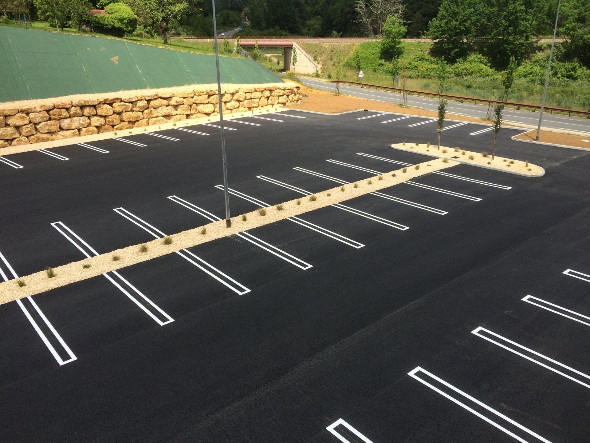 Parking vide avec asphalte noir neuf et marquages ​​blancs des places de stationnement, près d'un mur de pierre et d'une colline herbeuse.