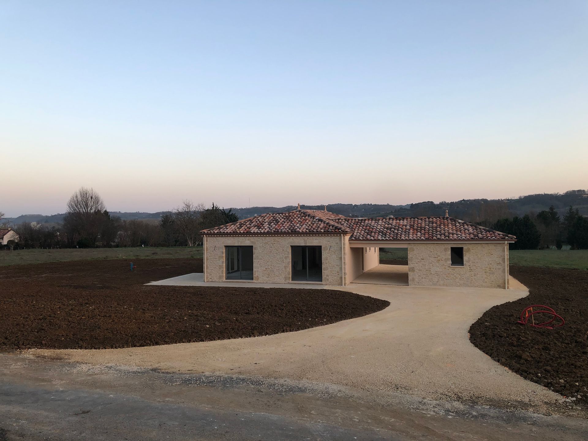 Petite maison moderne en construction sur une allée de gravier incurvée, entourée de terre nue au crépuscule