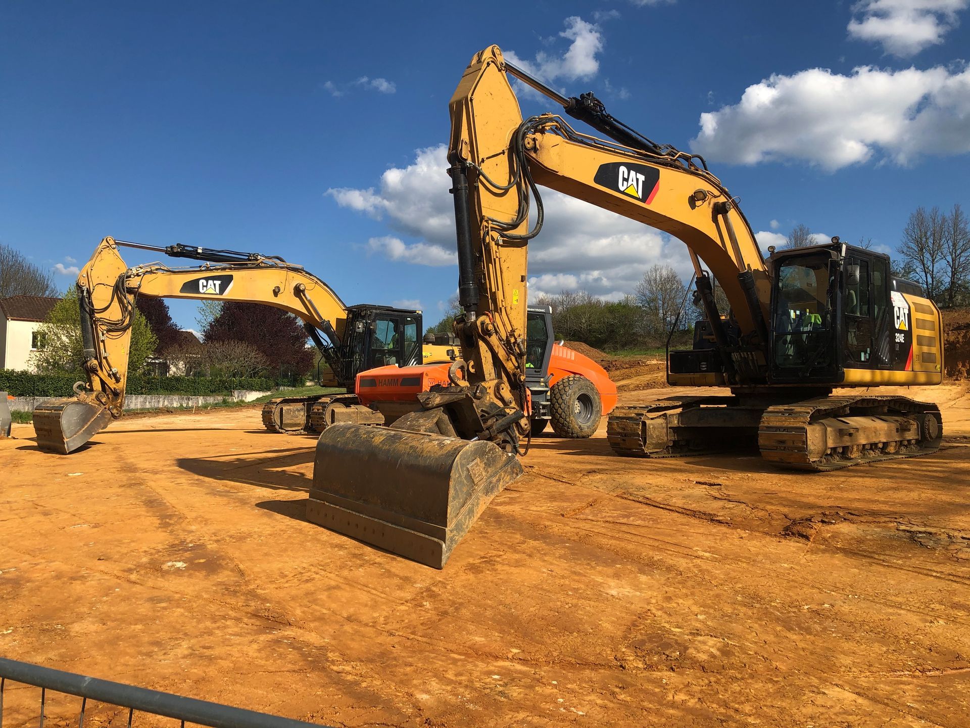 Deux excavatrices jaunes sur un terrain vague sous un ciel bleu, l'une d'elles avec le godet levé à l'avant.