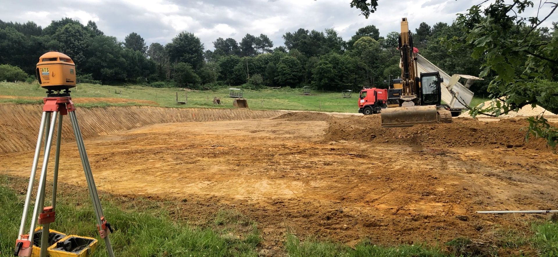 Trépied de topographie dans un champ boueux, avec une excavatrice et un tracteur orange en arrière-plan.