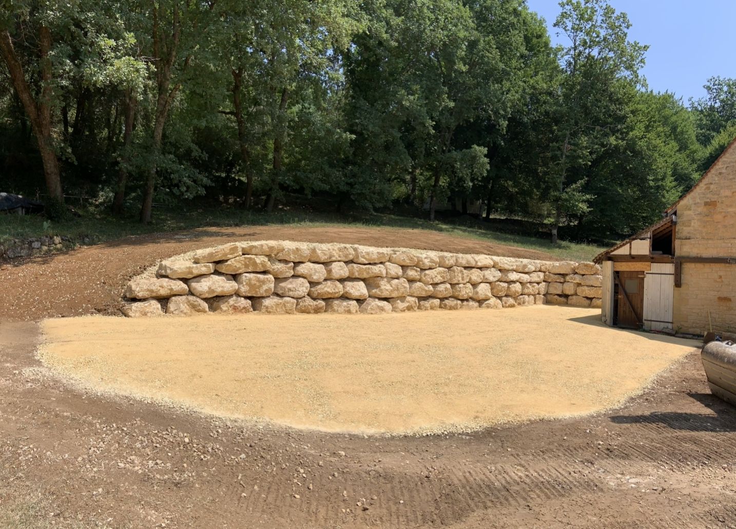 Mur de soutènement en pierre à côté d'une clairière en terre avec des arbres et un bâtiment en bordure d'une zone boisée