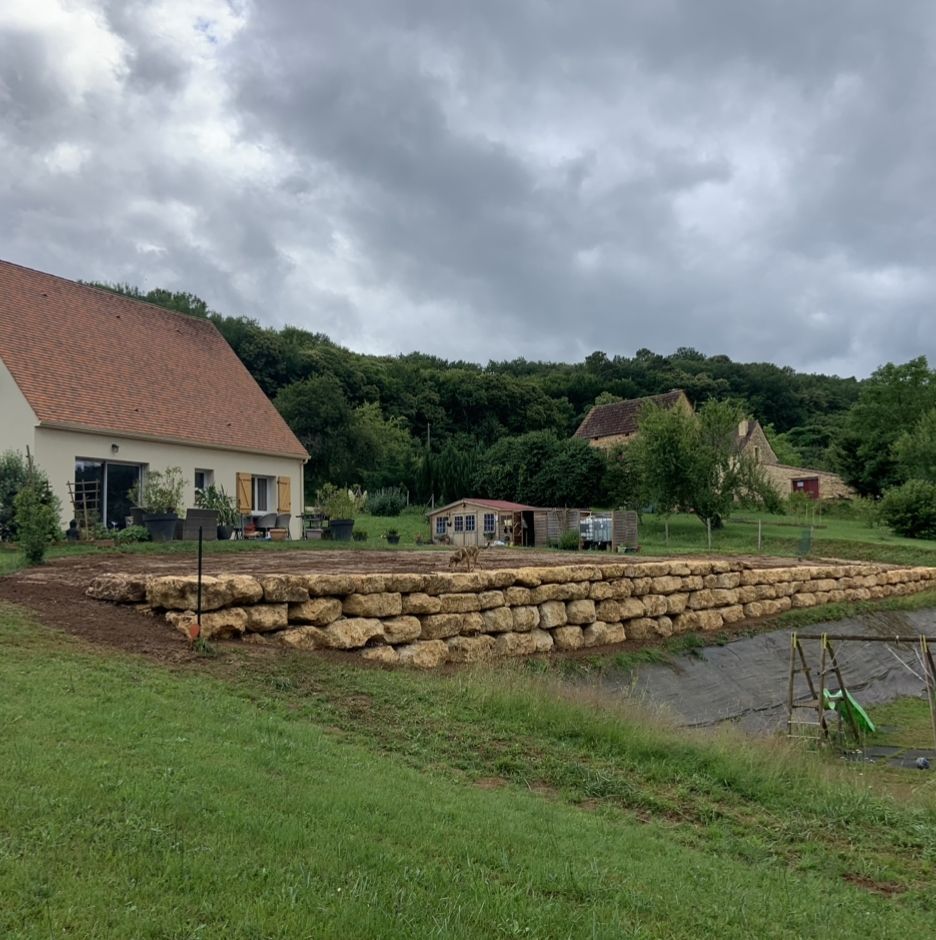 Maison rurale au toit rouge, adossée à un mur de soutènement en pierre sous un ciel nuageux, avec une cour herbeuse et des arbres.