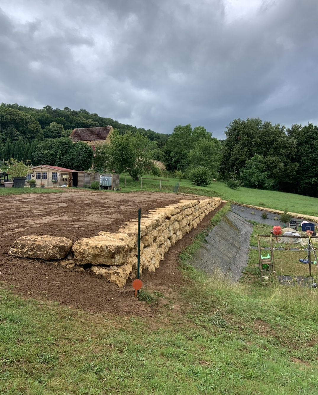 Mur de bottes de foin dans un champ herbeux sous un ciel nuageux, avec des arbres et un petit bâtiment en arrière-plan.