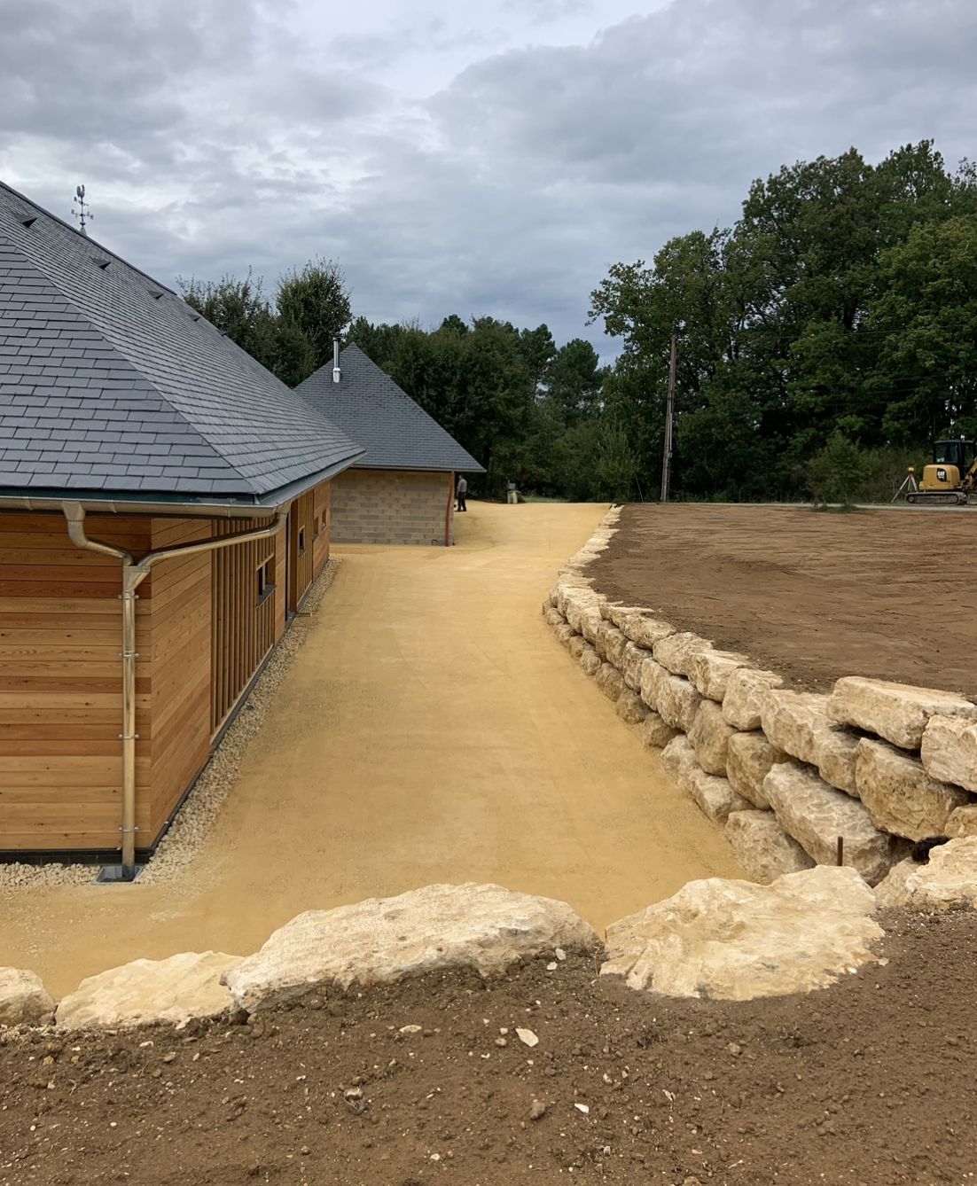 Chemin de gravier longeant un bâtiment en bois, bordé d'un aménagement paysager en pierres empilées sous un ciel nuageux.