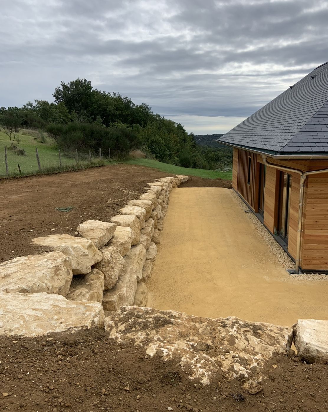 Chemin de gravier longeant une cabane en bois, bordé de pierres, traversant un flanc de colline rurale sous un ciel nuageux.