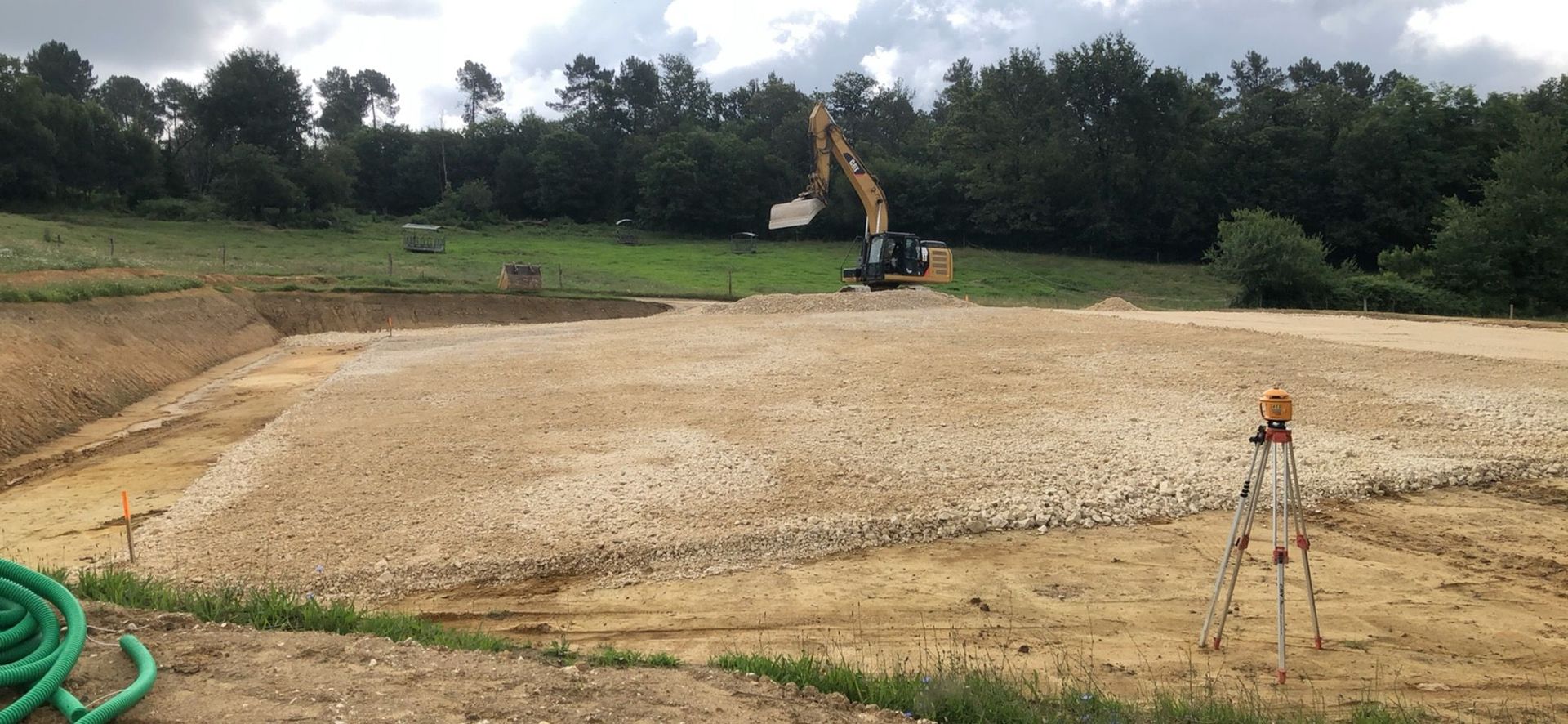 Un bulldozer et une excavatrice nivellent un champ de terre bordé d'une rangée d'arbres verdoyants sous un ciel nuageux.