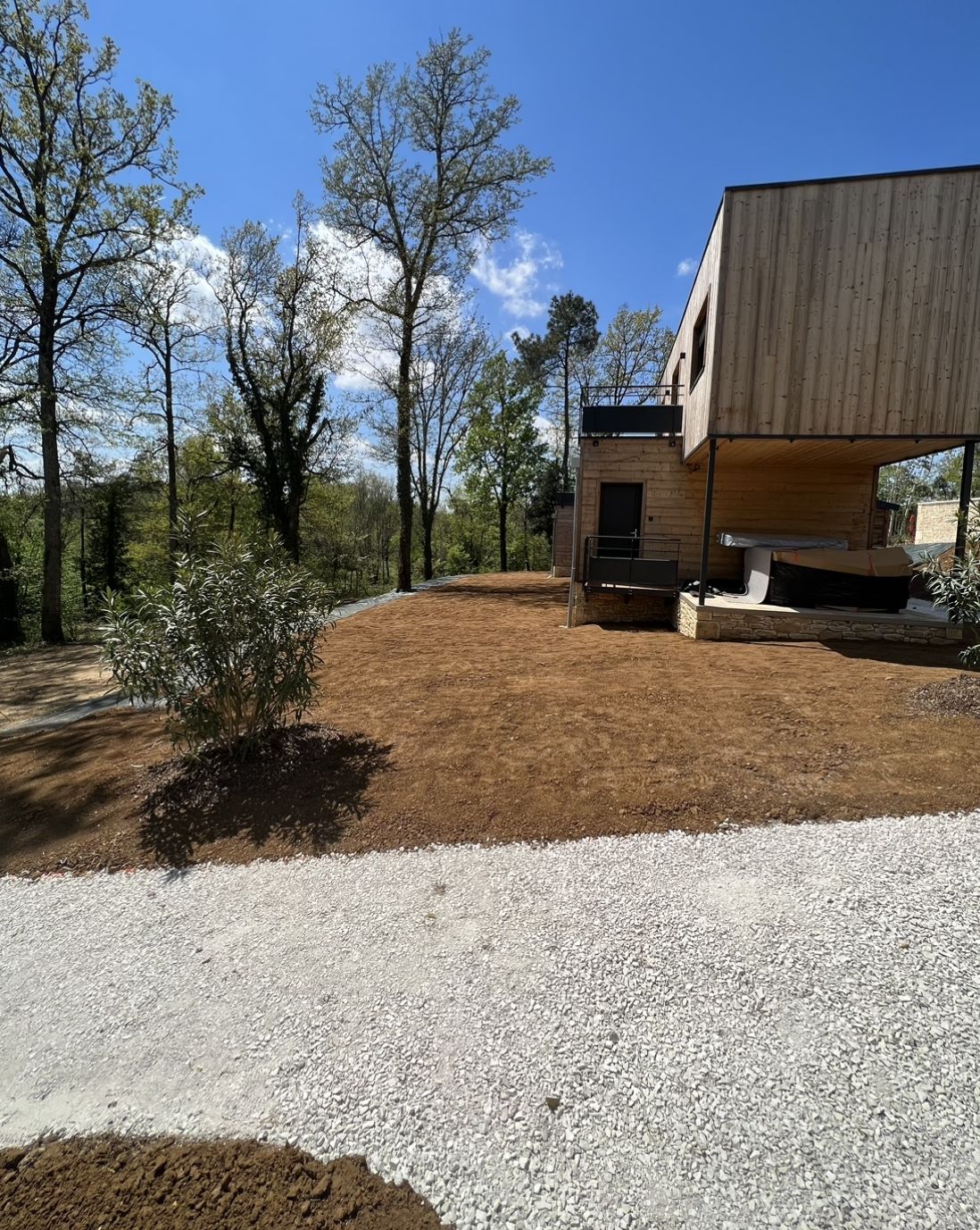 Maison moderne avec bardage en bois, située à côté d'une cour recouverte de gravier et de paillis, d'arbres et d'un ciel bleu.