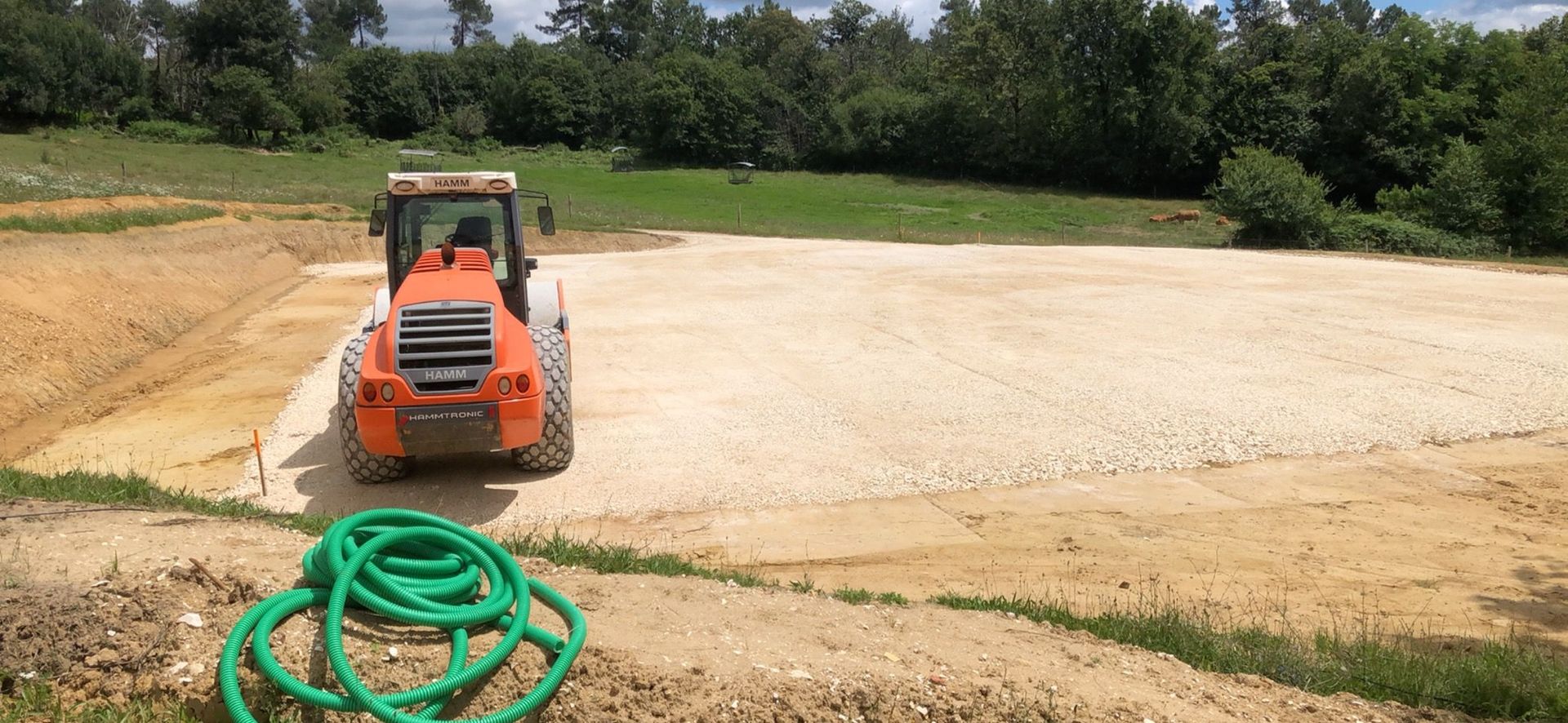 Une chargeuse compacte orange nivelle de la terre sur un chantier, avec un tuyau vert enroulé au premier plan.