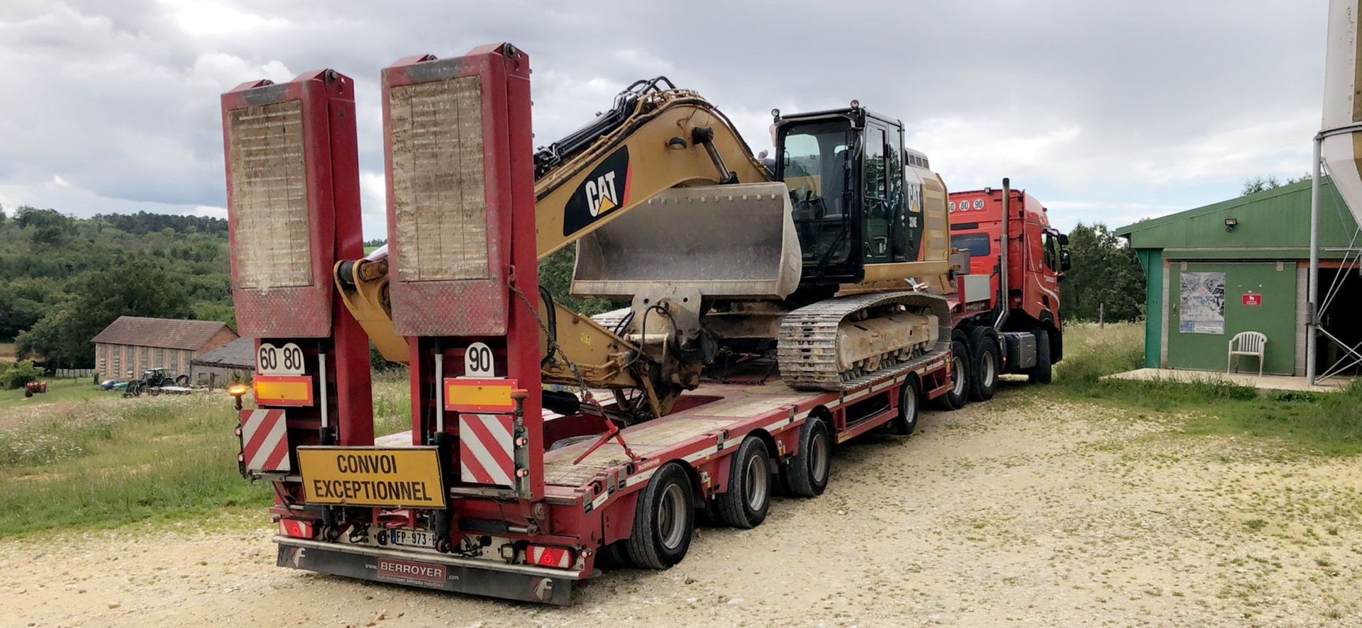 Une lourde pelle mécanique est chargée sur une remorque surbaissée rouge dans une cour gravillonnée, à proximité de camions et d'un hangar vert.