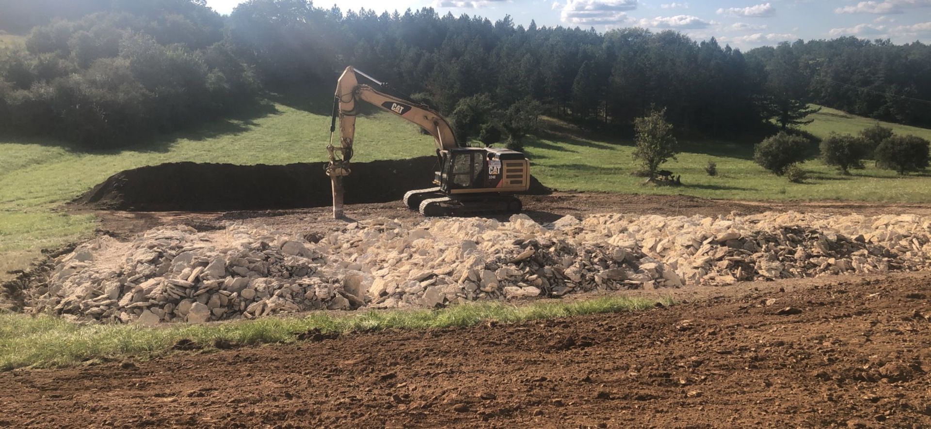 Une excavatrice creuse sur un terrain vague avec un monticule de terre, entouré de collines verdoyantes et d'arbres.