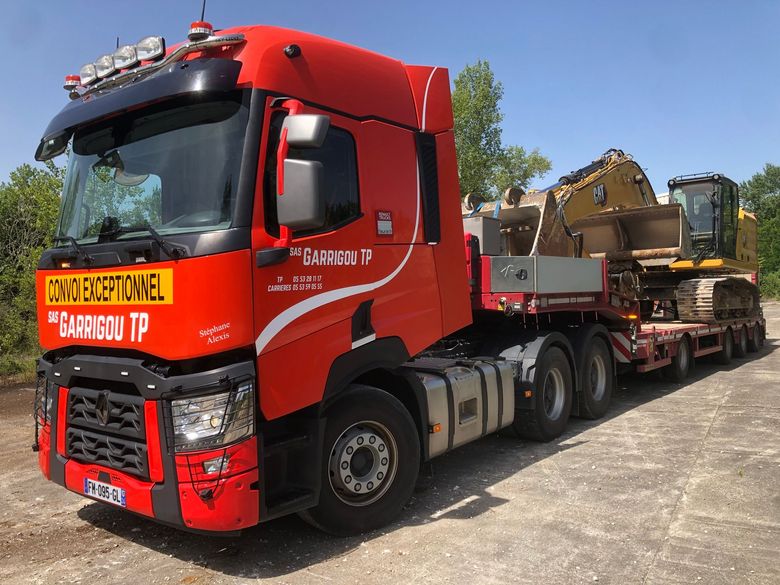 Un semi-remorque rouge tracte une remorque surbaissée chargée d'engins de chantier lourds sur une route ensoleillée.