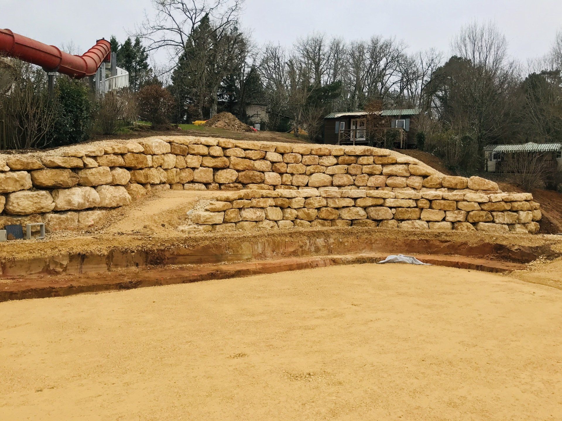 Mur de soutènement en pierre à plusieurs niveaux jouxtant une zone sablonneuse dans un cadre extérieur ressemblant à un parc.
