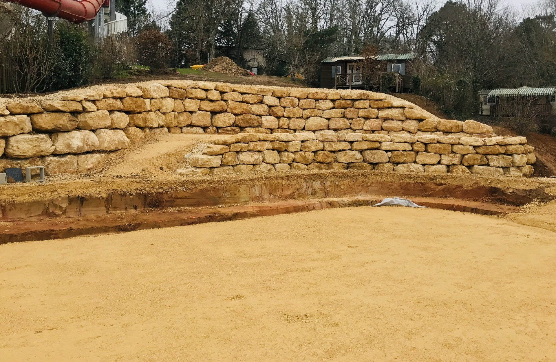 Mur de soutènement en pierre avec gradins bordant une zone extérieure sablonneuse et des arbres en arrière-plan