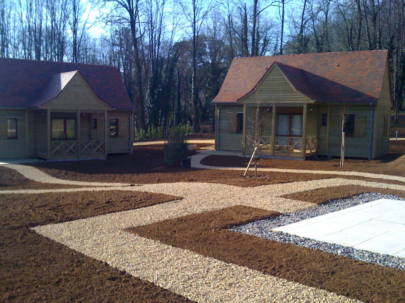Deux petites cabanes avec vérandas dans une zone boisée, avec un chemin de gravier et des arbres dénudés à proximité.